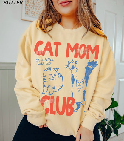 Beige hoodie with 'Cat Mom Club' text and cat illustrations worn by a person.