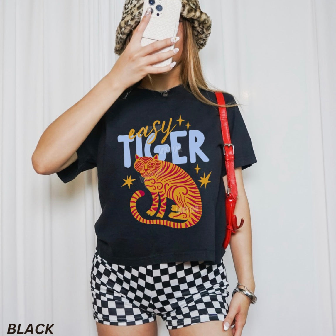 Person wearing a black t-shirt with a tiger graphic and 'easy Tiger' text, checkered shorts, and a red bag.