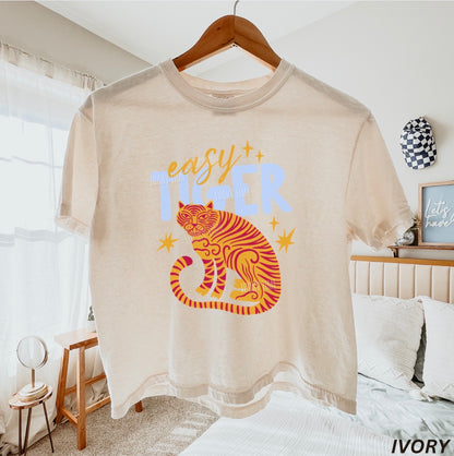 Beige t-shirt with tiger graphic and 'easy tiger' text on a hanger in a bedroom.