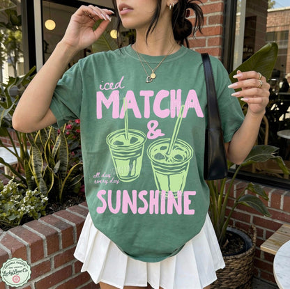 Person wearing a green t-shirt with 'iced matcha & sunshine' text and graphics outdoors.