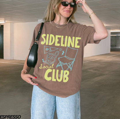 Person wearing a brown t-shirt with 'Sideline Social Club' text and graphics in an indoor setting.