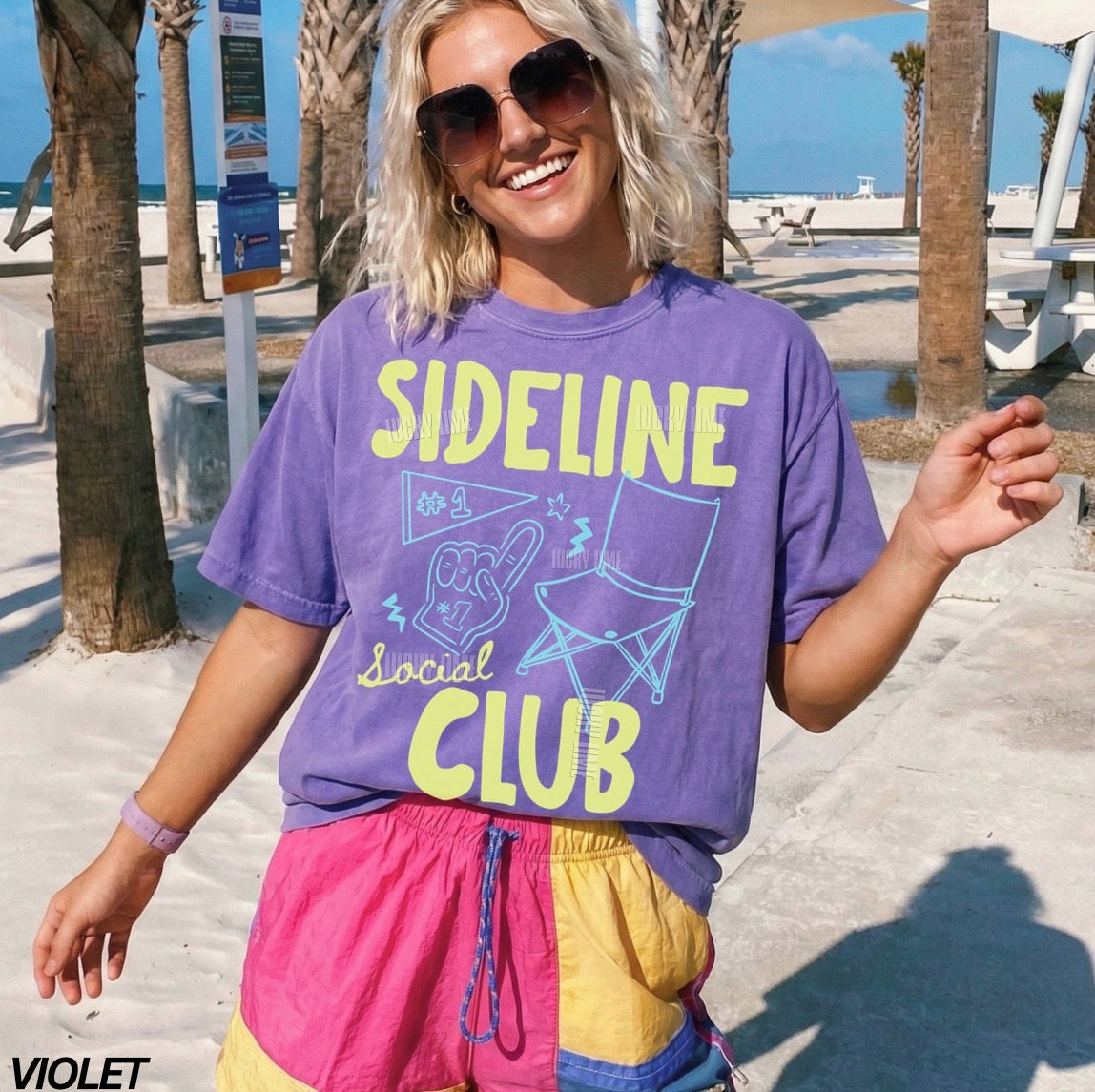 Person wearing a purple 'Sideline Social Club' t-shirt with sunglasses and colorful shorts on a beach.