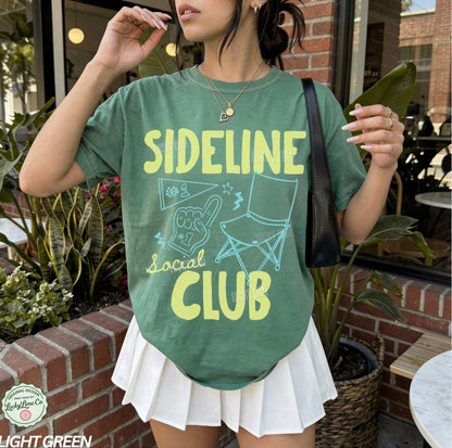 Person wearing a green t-shirt with 'Sideline Social Club' text and graphics outdoors.