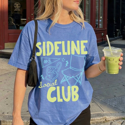 Person wearing a blue 'Sideline Social Club' t-shirt holding a drink on a sidewalk.