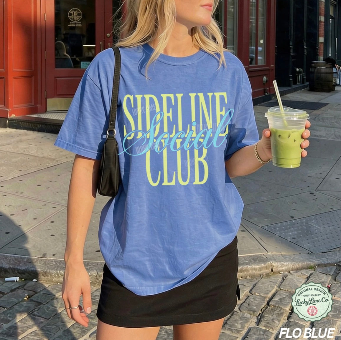 Woman wearing a blue 'Sideline Social Club' t-shirt holding a drink on a street.