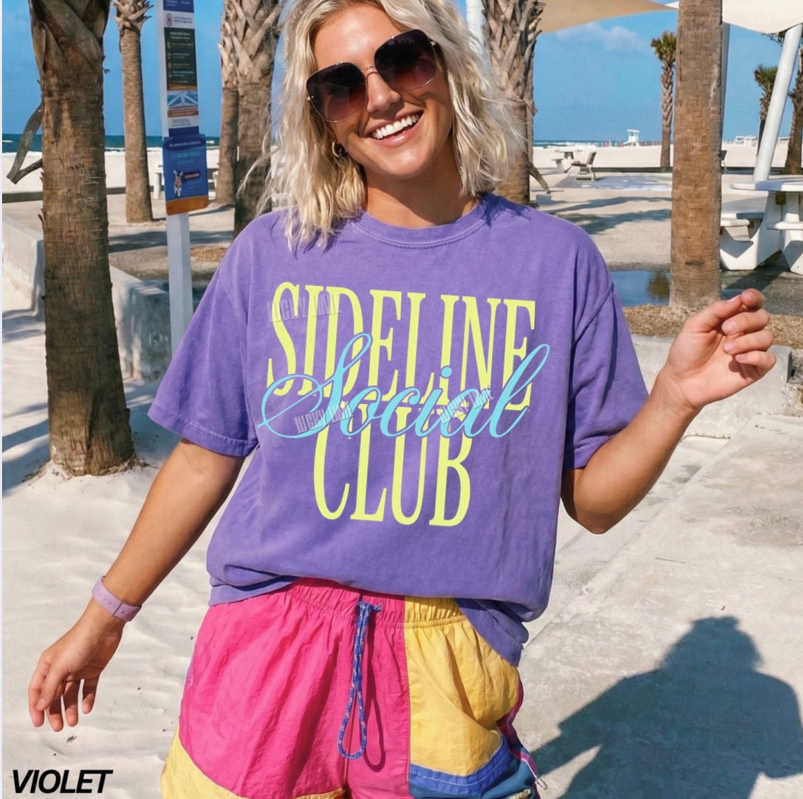 Woman wearing a purple 'Sideline Social Club' t-shirt and colorful shorts on a beach.