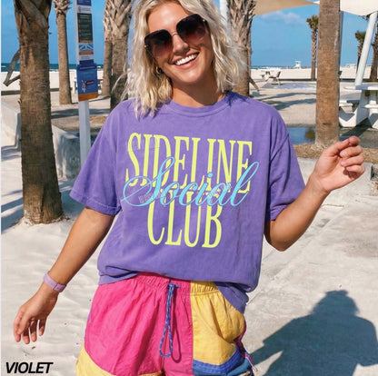 Woman wearing a purple 'Sideline Social Club' t-shirt and colorful shorts on a beach.