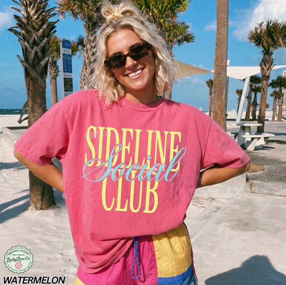Woman wearing a pink 'Sideline Social Club' t-shirt with palm trees and a beach in the background.