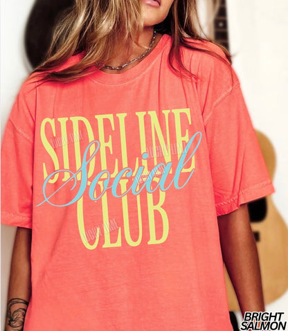 Person wearing a coral t-shirt with 'Sideline Social Club' text, blurred background