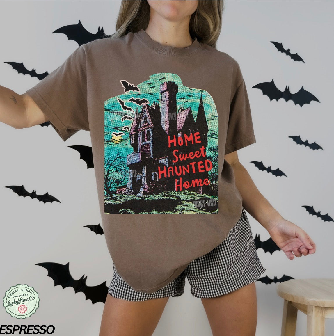 Person wearing a brown t-shirt with a Halloween-themed design featuring a haunted house, surrounded by bats on a light gray background.