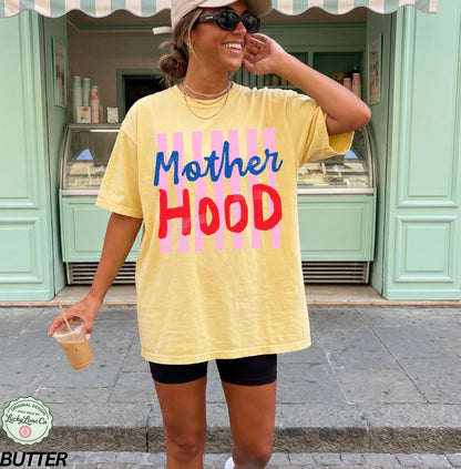 Woman wearing a yellow t-shirt with 'Mother Hood' text, standing in front of a green building.