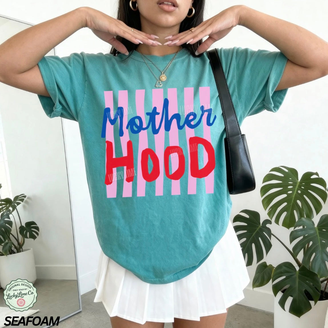 Person wearing a teal t-shirt with 'Mother Hood' text and pink stripes, standing indoors with plants.