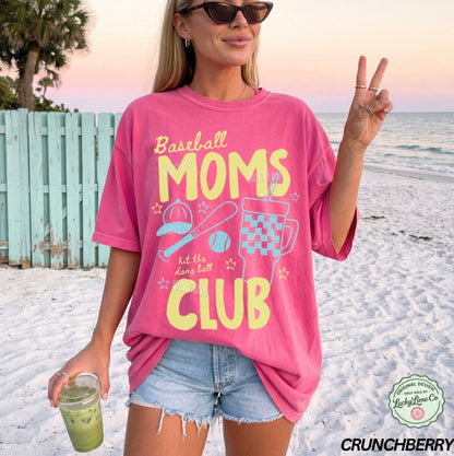 Woman on a beach wearing a pink t-shirt with 'Baseball Moms Club' text, holding a drink.
