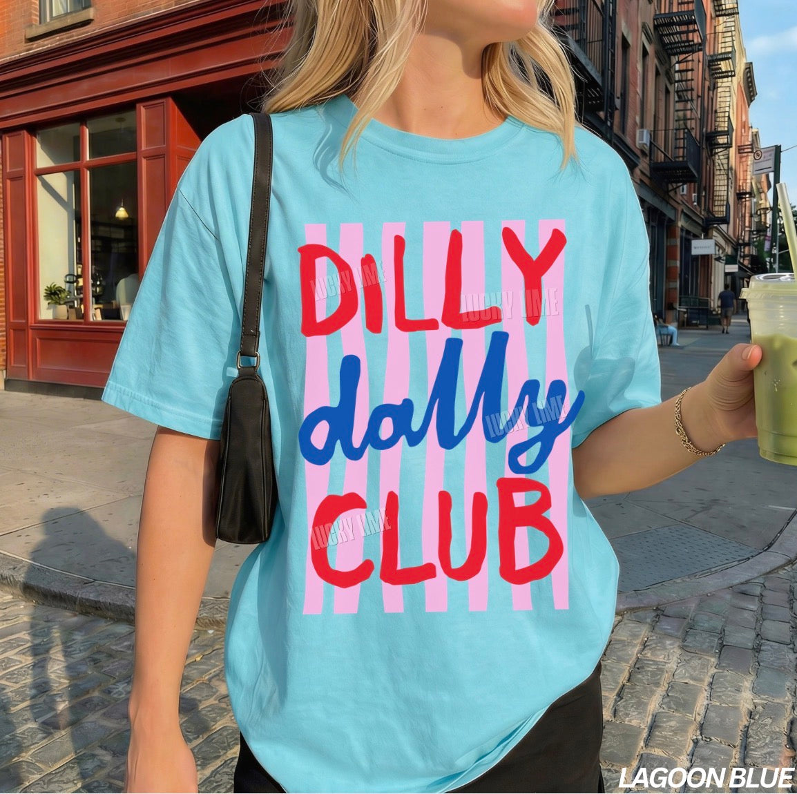 Person wearing a light blue t-shirt with 'Dilly Dally Club' text on a city street.