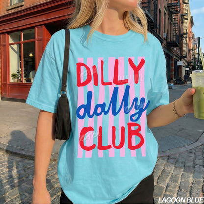 Person wearing a light blue t-shirt with 'Dilly Dally Club' text on a city street.