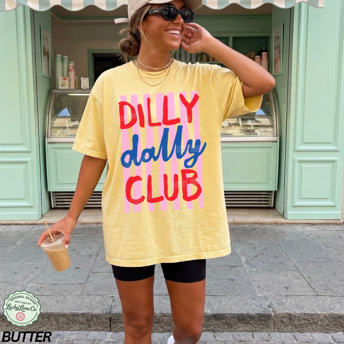 Woman wearing a yellow t-shirt with 'Dilly Dally Club' text, standing in front of a green building.