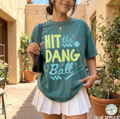 Woman wearing a dark green/blue  tshirt with the words “Hit the Dang ball” with baseball graphics