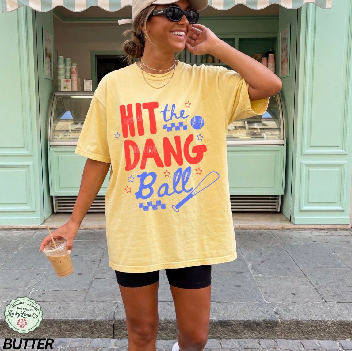 Woman wearing a yellow t-shirt with 'Hit the Dang Ball' text, standing in front of a green building.
