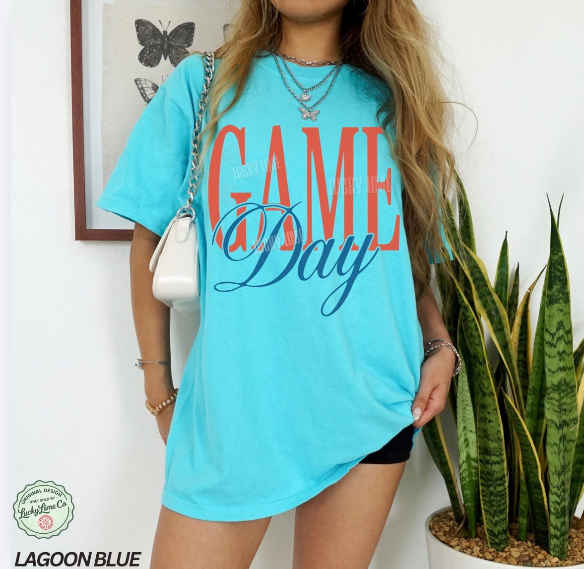 Person wearing a light blue 'Game Day' t-shirt with a plant and framed picture in the background.