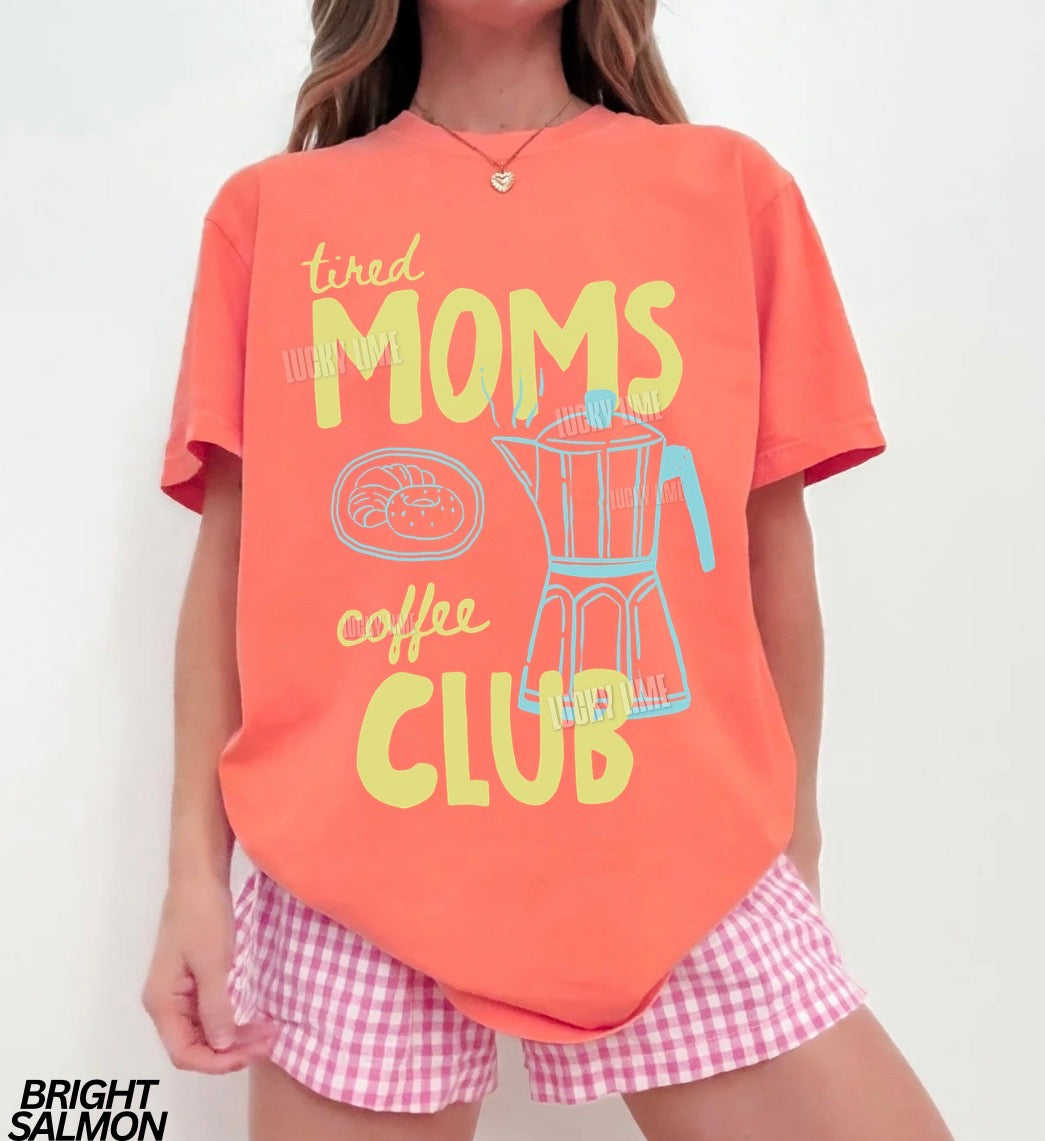 Person wearing a bright salmon t-shirt with 'tired moms coffee club' text and checkered shorts on a white background.