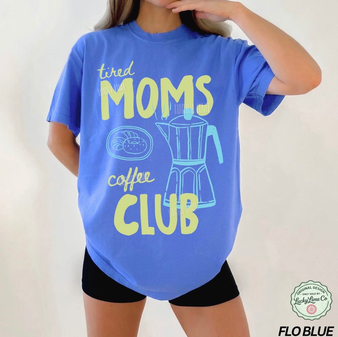 Person wearing a blue t-shirt with 'tired moms coffee club' text and graphic on a plain background