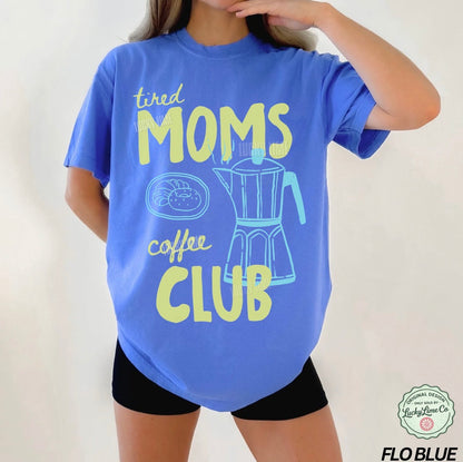 Person wearing a blue t-shirt with 'tired moms coffee club' text and graphic on a plain background