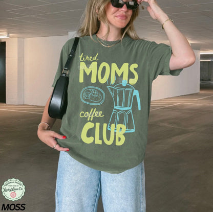 Woman wearing a green t-shirt with 'tired moms coffee club' text and design, holding sunglasses.