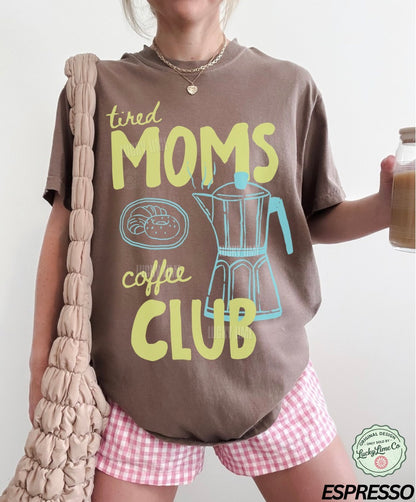 Person wearing a brown t-shirt with 'tired moms coffee club' text and graphics, holding a jar of coffee.