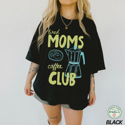 Person wearing a black t-shirt with 'tired moms coffee club' text and graphics on a plain background