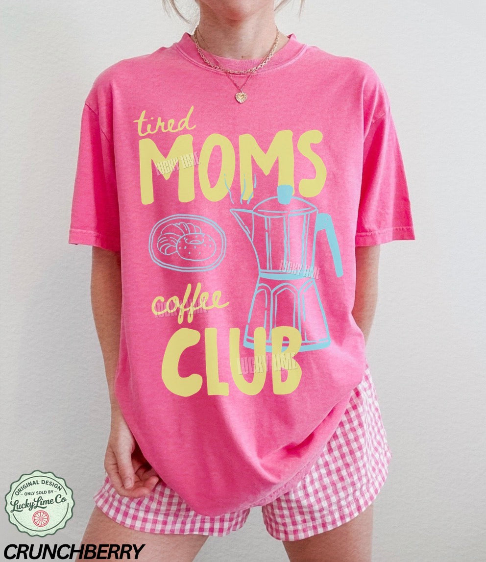 Person wearing a pink 'tired moms coffee club' shirt with checkered shorts on a plain background