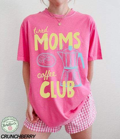 Person wearing a pink 'tired moms coffee club' shirt with checkered shorts on a plain background
