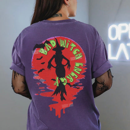 Person wearing a purple t-shirt with a graphic design on the back, standing in front of a neon sign.