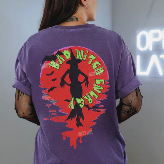 Person wearing a purple t-shirt with a graphic design on the back, standing in front of a neon sign.