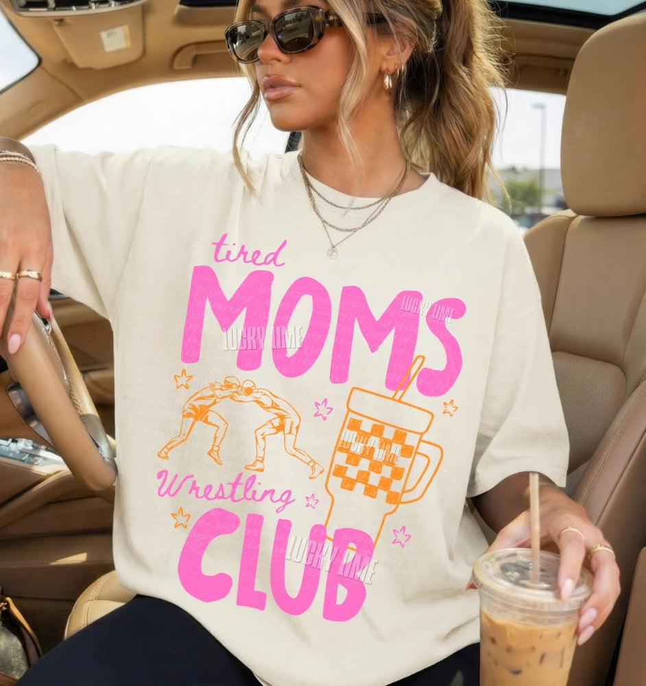 Woman sitting in a car wearing a t-shirt with 'tired moms wrestling club' text and graphics.