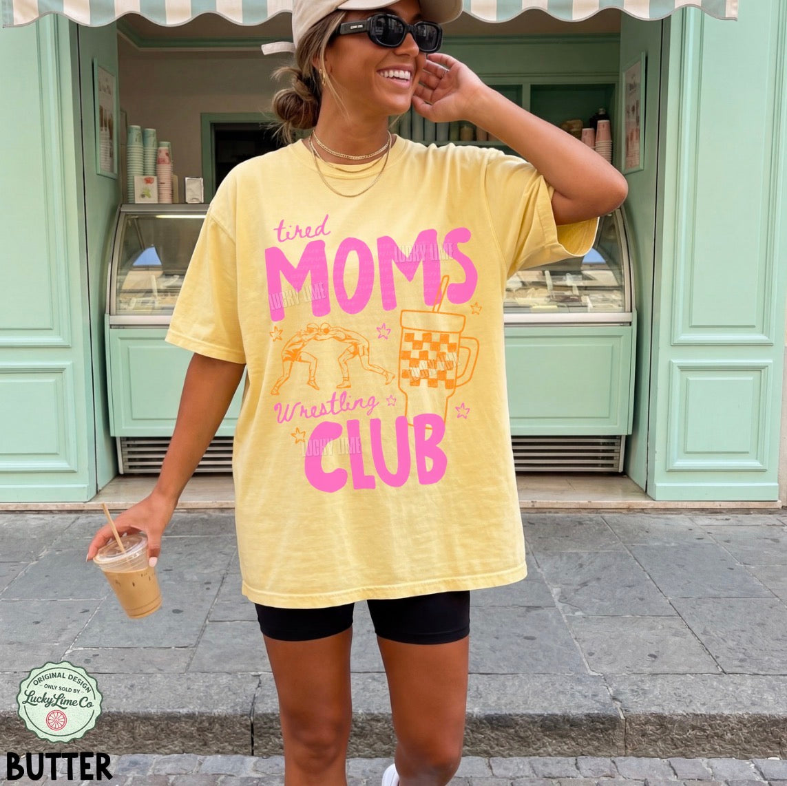 Woman wearing a yellow t-shirt with pink text and graphics, standing in front of a green building.