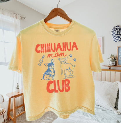 Yellow t-shirt with 'Chihuahua mom club' text and graphics on a hanger in a bedroom.