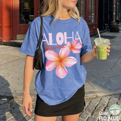 Person wearing a blue 'Aloha Hawaii' t-shirt with floral design, holding a green drink.