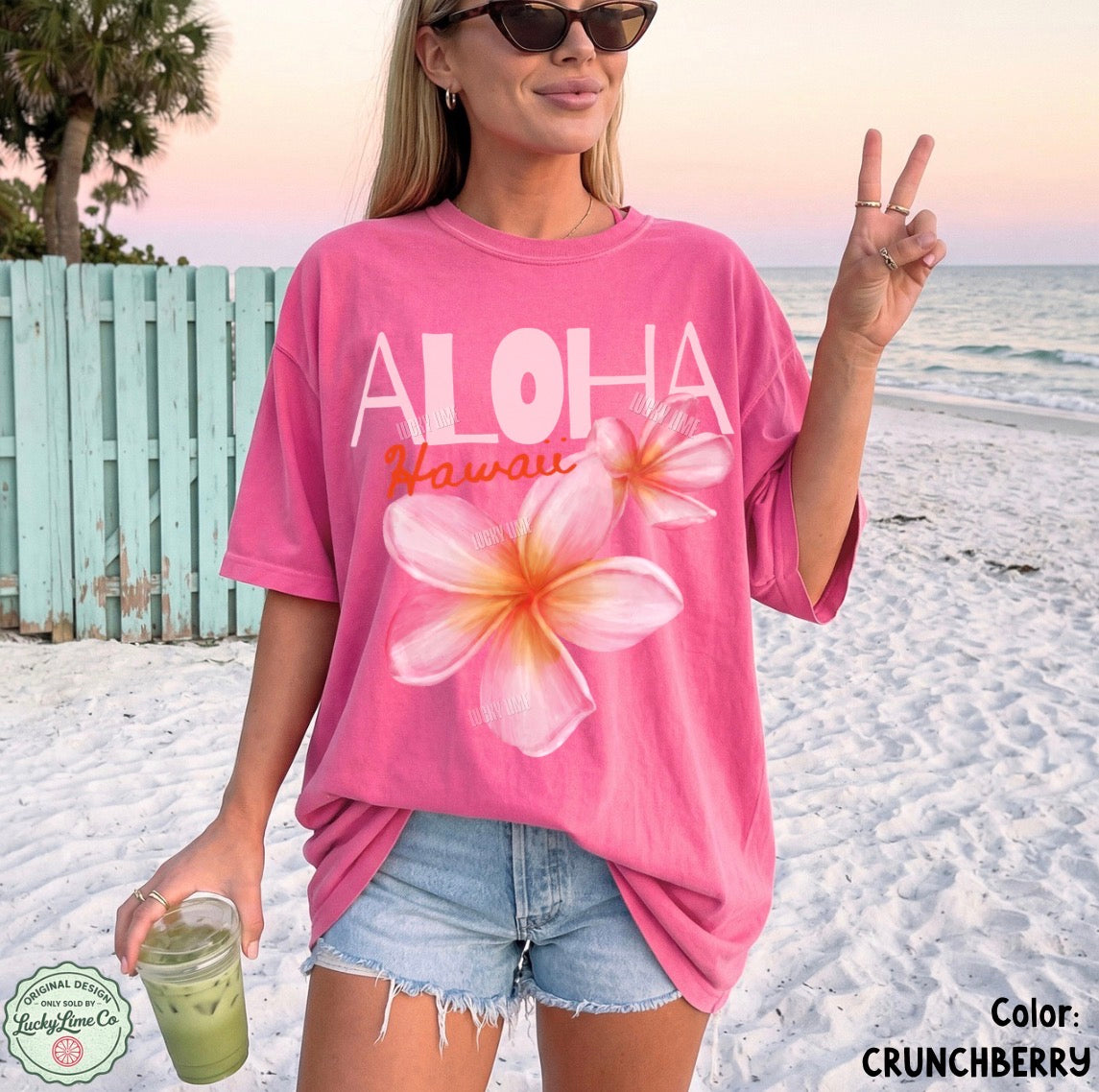 Woman on a beach wearing a pink 'Aloha Hawaii' t-shirt with floral design, holding a drink.