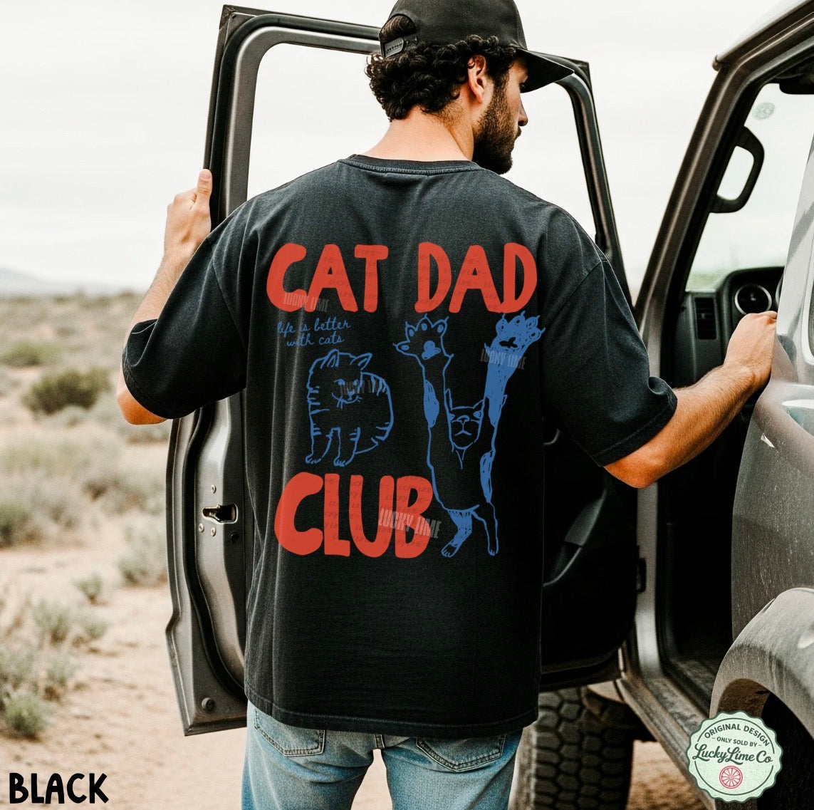 Man wearing a black t-shirt with 'Cat Dad Club' text and graphics, standing next to an open car door in a desert setting.