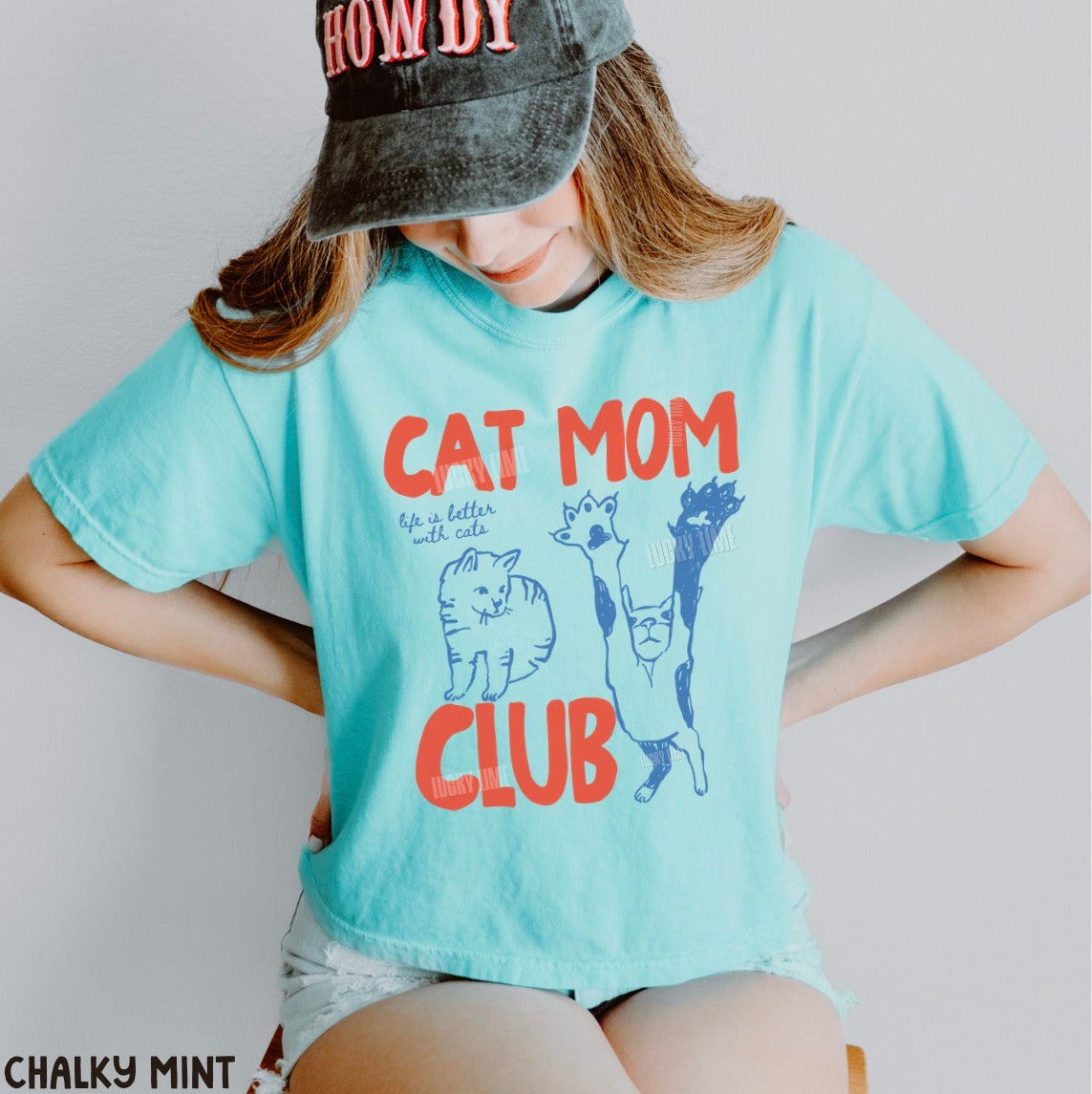 Person wearing a light blue t-shirt with 'Cat Mom Club' text and graphics, standing against a plain background.