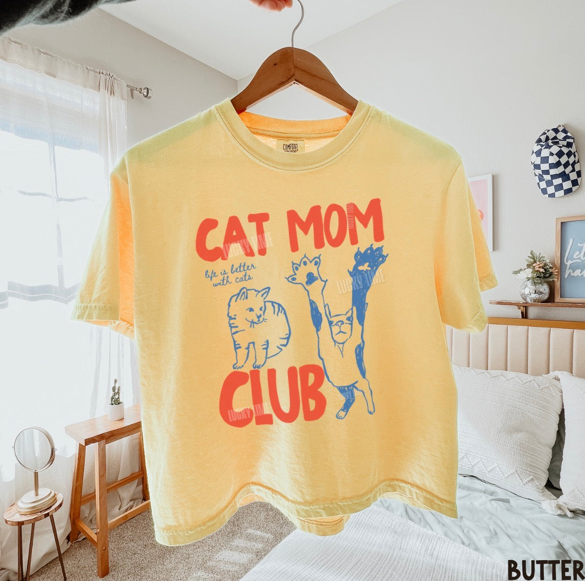 Yellow t-shirt with 'Cat Mom Club' text and graphics on a hanger in a bedroom.