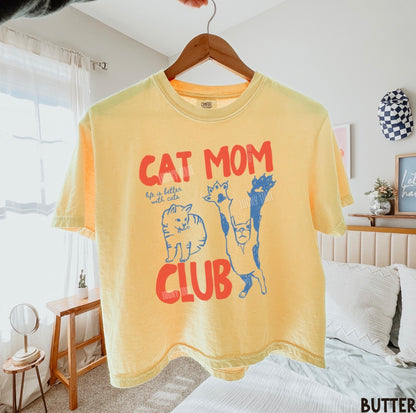 Yellow t-shirt with 'Cat Mom Club' text and graphics on a hanger in a bedroom.