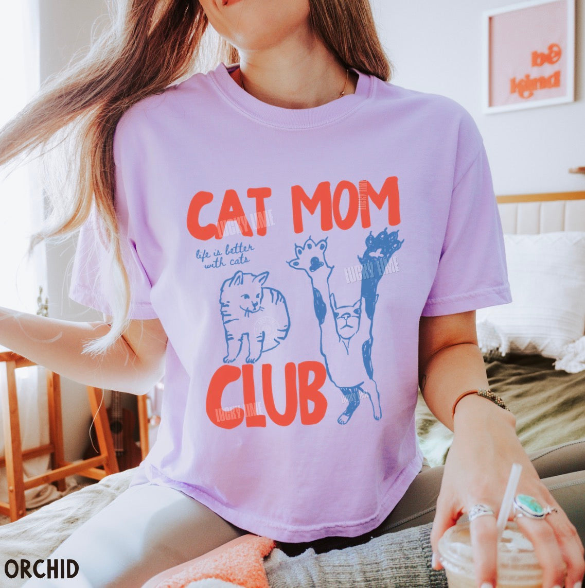 Person wearing a purple t-shirt with 'Cat Mom Club' design, sitting indoors.