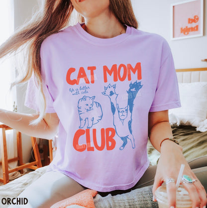 Person wearing a purple t-shirt with 'Cat Mom Club' design, sitting indoors.