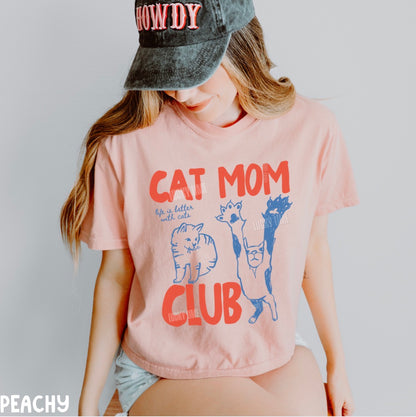 Person wearing a pink t-shirt with 'Cat Mom Club' design and a cap.