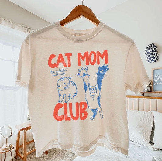 Beige t-shirt with 'Cat Mom Club' text and cat illustrations on a hanger in a bedroom.