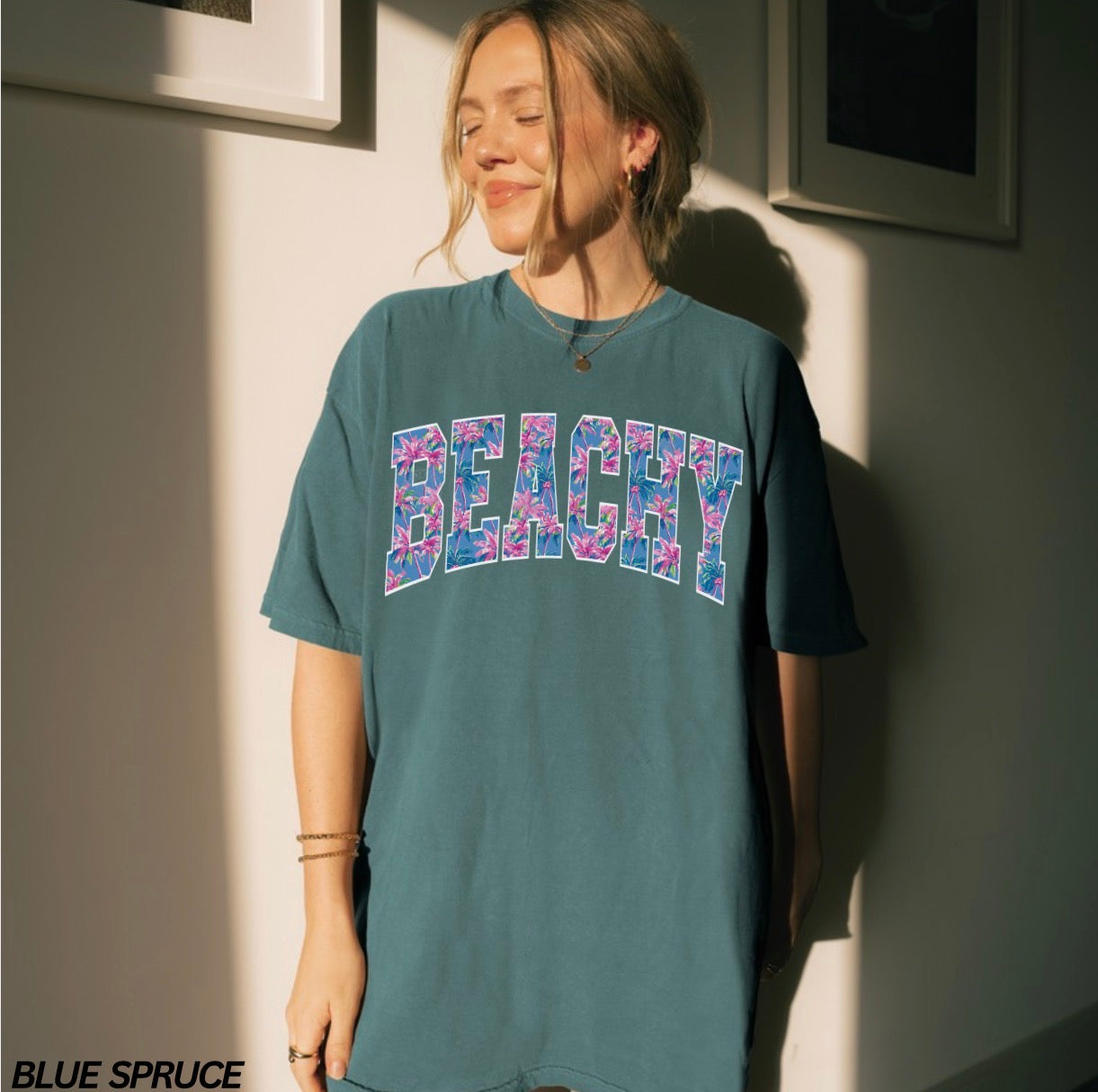 Person wearing a teal t-shirt with 'BEACHY' printed in colorful letters against a neutral wall.