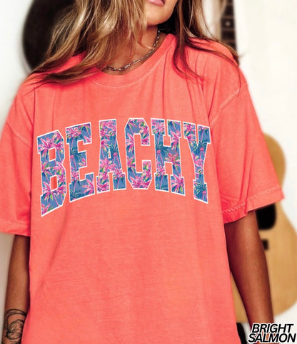 Coral t-shirt with 'BEACHY' text in floral letters worn by a person.