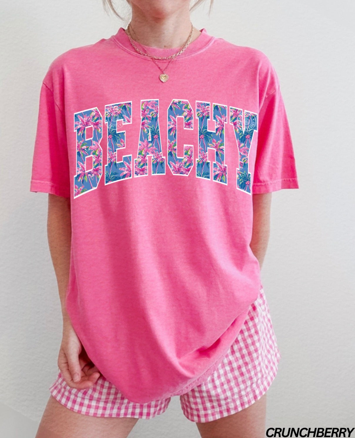 Person wearing a pink 'BEACHY' t-shirt with floral letters and checkered shorts on a plain background.