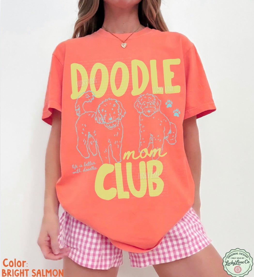 Person wearing a bright salmon t-shirt with 'Doodle Mom Club' text and checkered shorts on a white background.